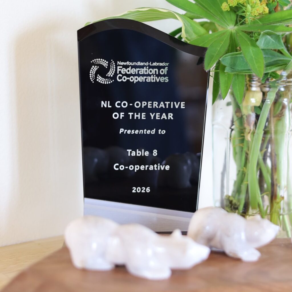 The award is vertical black reflective material like stone. The top of the award is carved into a wave shape. The award is sitting on a table beside a beautiful arrangement of wild newfoundland flowers. Engraved on the award says "Newfoundland-Labrador Federation of Co-operatives NL Co-operative of the year presented to Table 8 Co-operative 2026"